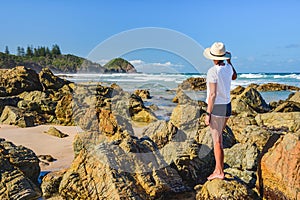 Australian coast, a young woman in a hat, T-shirt and shorts stands on the rocks on the beach and looks at the seaside landscape