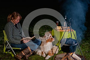 Australian Shepherd dog is lying by the campfire, the kettle with food is cooking and steaming, The woman is reading a