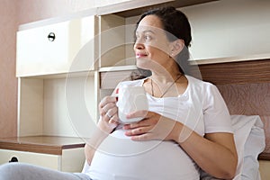 Authentic adult pregnant woman with cup of hot tea, dreamily looking away, experiencing happy moments of her pregnancy