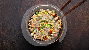 Authentic Chinese and Asian fried rice with egg and vegetables in ceramic brown bowl top view, dark rustic concrete