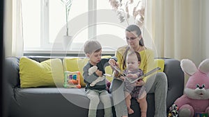 Authentic close up cute neo caucasian spectacled mom with 2 children boy and girl on gray yellow sofa reading book