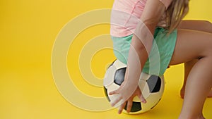 Authentic cute smiling preschool little girl sits on classic black white soccer ball look at camera on yellow background