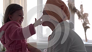 Authentic cute young woman applying face mask of baby girl. mom with 2 children daughters different ages in bathrobes