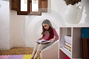 Authentic portrait erudite child girl, elementary age schoolgirl reading a book in her room. Nearby bookshelf with books