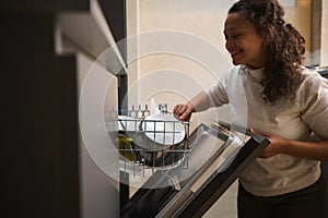 Authentic portrait of a happy smiling woman using a modern dishwasher in the kitchen. Housekeeping. Household chores