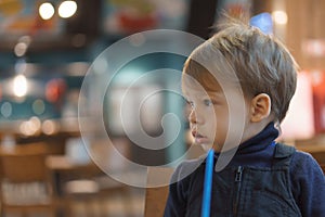 authentic portrait little handsome blond sad boy sitting in cafe in evening alone. Preschool toddler kid child eating