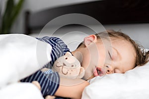 Authentic portrait sick cute caucasian little preschool baby boy in blue sleep with teddy bear on white bed. child