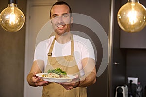 Authentic portrait of a smiling Caucasian man in beige chef's apron, holding a plate with Italian spaghetti pasta