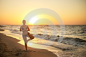 Authentic woman in swimsuit doing yoga vrikshasana on the beach in the morning. Real unretouched shape girl silhouette