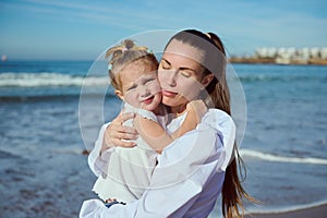 Authentic young happy affectionate mother gently hugs her adorable little child daughter, walking together on the beach