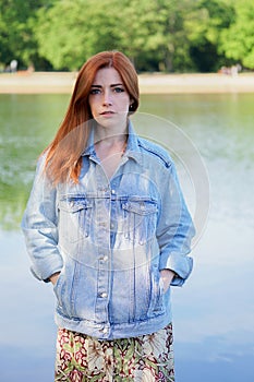 Authentic young woman wearing denim jacket over summer dress standing by lake
