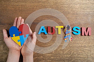 Autistic boy hands holding heart shapeed puzzle with word autism and awarennes ribbon on wooden background. Autism