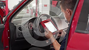 Auto electrician uses OBD2 scanner and tablet to diagnose car in garage. Technician reads fault codes, checks vehicle