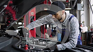 Auto mechanic examining broken car engine, standing near his car with open hood in a car repair shop