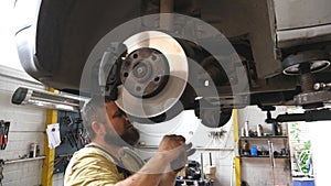 Auto mechanic fixing suspension of a car at garage. Professional bearded repairman in uniform working underneath a