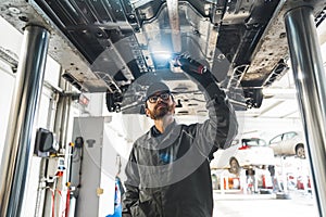 Auto mechanic with LED work light inspecting car chassis standing under it