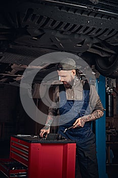 Auto mechanic in a uniform, working on a workbench while standing under lifting car in a repair garage.