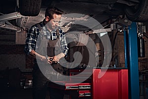 Auto mechanic in a uniform, working on a workbench while standing under lifting car in a repair garage.