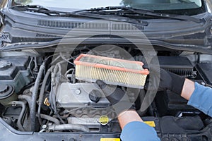 Auto mechanic wearing protective work gloves holding used air filter above a car engine