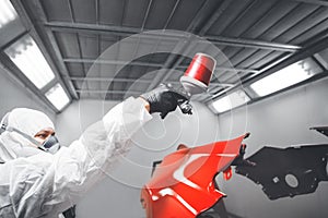 Auto mechanic worker painting car element with spray gun in a paint chamber during repair work.