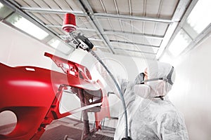 Auto mechanic worker painting car element with spray gun in a paint chamber during repair work.