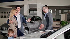 auto sale center, young male manager telling happy male and female buyers about new automobile in car showroom