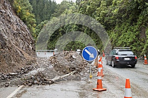 Automobile car driving past rockflall, rocks, landslide debris, traffic cones and warning signs on roadway highway. Dangerous road