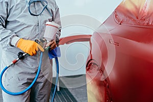 Automobile repairman worker painting a red car in a paint chamber during repair work.