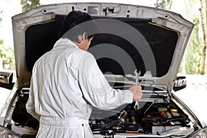 Automotive mechanic in uniform with wrench diagnosing engine under hood of car at the repair garage.