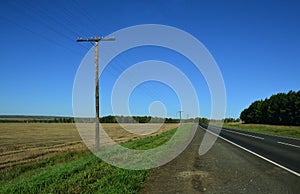 The automotive side of the paved road. Telegraphic wooden poles and a sloping field along the road.