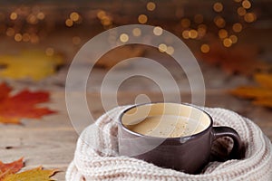 Autumn background from cup of cocoa or coffee in knitted scarf on wooden table decorated with fall leaves. Cozy hot drink.