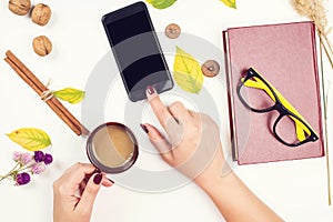 Autumn background with mobile phone, autumn leaves, vintage book on white desk. Top view. Woman hand holding cup of coffee. Girl