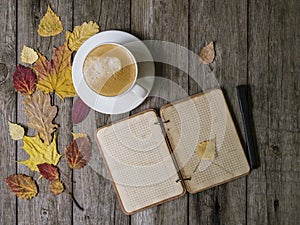 Autumn background on a wooden table with autumn dry colored leaves, white cup of coffee and a notepad with a pencil