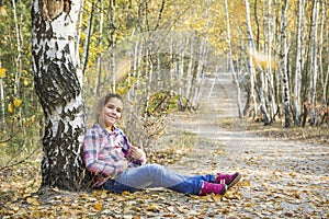 In the autumn birch forest, a little girl sits under a tree near the path