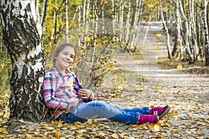 In the autumn birch forest, a little girl sits under a tree near the path