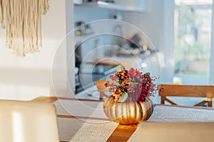 Autumn bouquet of bright artificial flowers in a golden pumpkin vase on a kitchen table counter with white modern