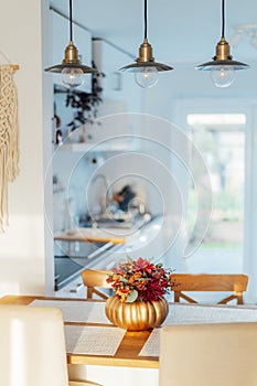 Autumn bouquet of bright artificial flowers in a golden pumpkin vase on kitchen table counter with white modern kitchen