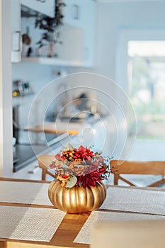 Autumn bouquet of bright artificial flowers in a golden pumpkin vase on kitchen table counter with white modern kitchen