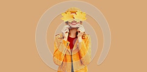 Autumn color style outfit, portrait of beautiful smiling young woman covering her eyes with yellow maple leaves wearing orange