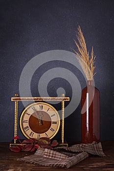 Autumn composition, brown bottle with dry leaves, vintage golden clock, fabric napkin on deep grey background. Vertical shot, copy