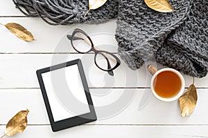 Autumn composition. Feminine desk table with knitted scarf, tea cup, glasses, tablet ebook with blank screen, fall leaves on
