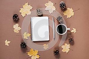 Autumn composition. Golden maple leaves, coffee cup, bumps, notebook and pen on brown background.