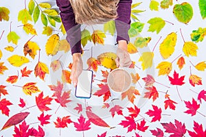 Autumn composition: top view hands holding cup of coffee and smatrphone with blank screen on fallen leaves gradient colorful rainb