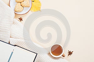 Autumn composition. White knitted plaid, cup of tea, book with empty pages, cookies, fallen leaves on pastel beige background.