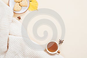 Autumn composition. White knitted plaid, cup of tea, cookies, fallen leaves on pastel beige background. Flat lay, top view, copy