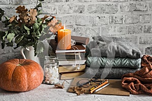 Autumn cozy home still life. Pumpkin, stack of books, burning candle, pile of winter autumn sweaters, jar of marshmallows on the