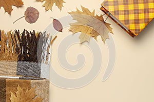 Autumn, fall concept. Checked orange gift paper bag, warm cozy blanket plaid, autumn dried leaves on pastel beige background. Flat