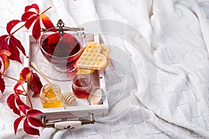 Autumn fall concept with knitted blanket and hot tea with waffer, jam, honey on wooden tray on te bed, autumn leaf and