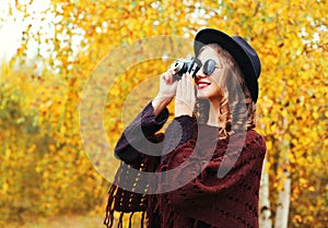 Autumn fashion smiling woman with retro vintage camera wearing black hat sunglasses and knitted poncho over sunny yellow leaves