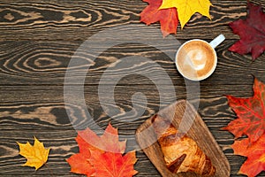 Autumn flat lay with a cup of coffee, homemade croissant and colorful maple leaves on the wooden background. Morning ritual.